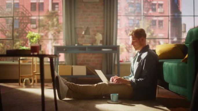 一个英俊的年轻成年男性坐在地板上，在阳光明媚的阁楼客厅使用笔记本电脑的肖像。成功的人在家在光明公寓工