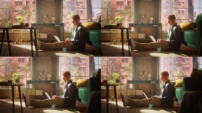 一个英俊的年轻成年男性坐在地板上，在阳光明媚的阁楼客厅使用笔记本电脑的肖像。成功的人在家在光明公寓工
