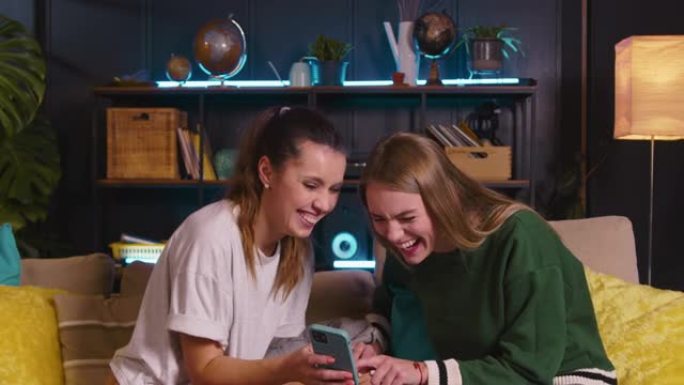 两个快乐的年轻美丽的高加索女性朋友在智能手机上观看有趣的视频，在舒适的家中在沙发上大笑。