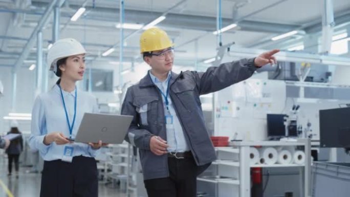 两名年轻的亚洲重工业工程师戴着安全帽，带着笔记本电脑在工厂里散步。两名制造业员工在研发机构工作。中等