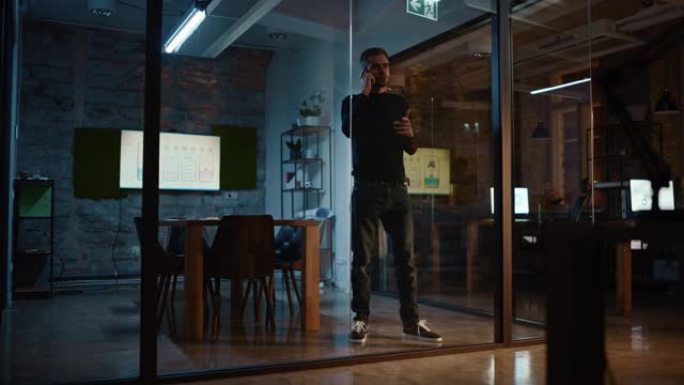 英俊的白人男性站在一家创意机构玻璃墙后面的会议室里打电话。项目经理谈智能手机上的商业策略