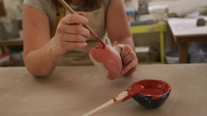 女性高加索陶工的中间部分穿着围裙，用刷子在陶器工作室涂漆锅
