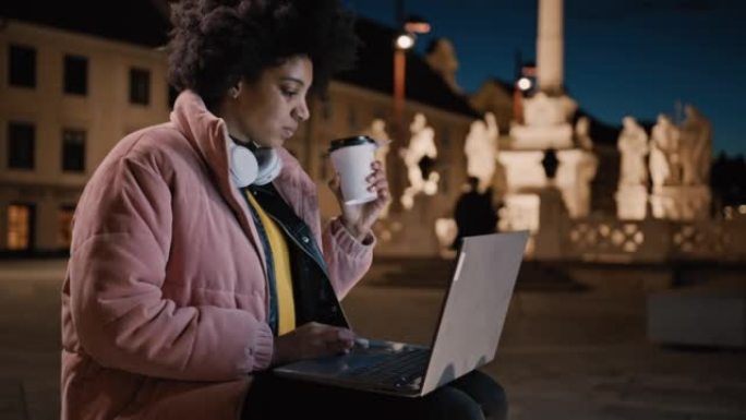 年轻女子坐在城市的长凳上，一边用笔记本电脑工作，一边喝咖啡