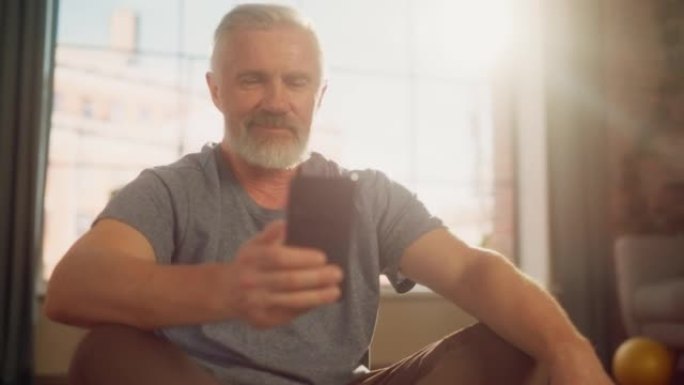 在阳光明媚的公寓里，一个健康的中年男子在晨练期间在锻炼之间使用智能手机的特写肖像。健康的生活方式，健