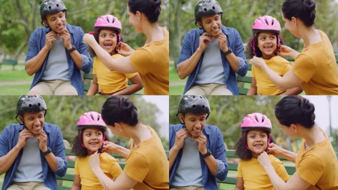 一个快乐的多元化家庭，坐在公园长椅上，给女儿戴上粉红色的头盔，然后在户外玩得开心。深情的父母检查孩子
