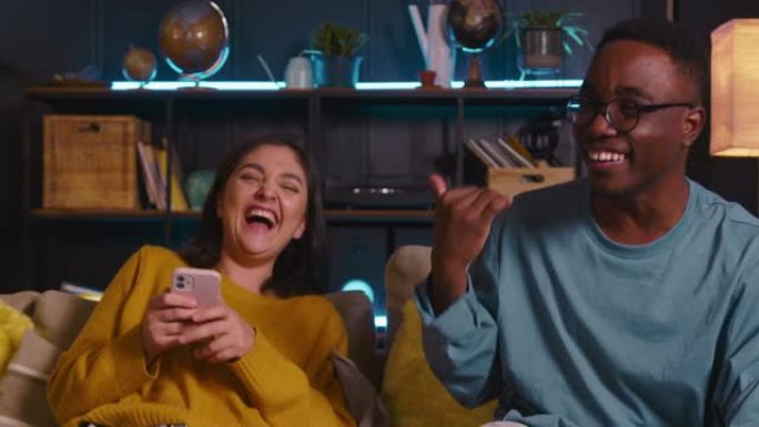 有趣快乐的年轻多民族夫妇使用手机浏览社交媒体，不能停止在家里一起大笑。