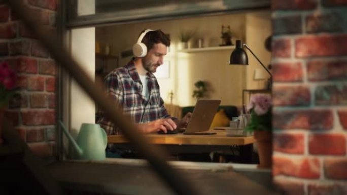英俊的白人男子坐在家庭办公室的桌子后面，在笔记本电脑上工作。自由企业家听音乐，做远程工作。从户外到窗
