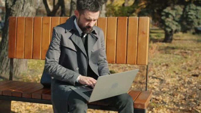 慢动作严肃的商人使用笔记本电脑坐在户外的长凳上