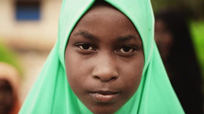 一个害羞的真实非洲女孩在镜头前微笑的特写肖像，背景中模糊的孩子在玩耍。黑人女性儿童对世界的未来和团结