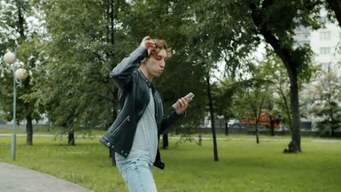 快乐的家伙在城市公园户外跳舞，独自戴着耳机和使用智能手机行走
