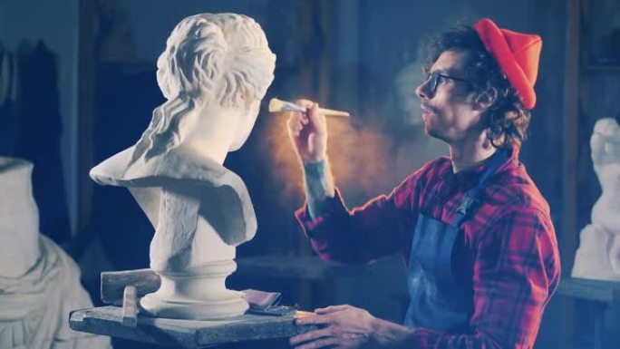 戴着眼镜和无檐小便帽的艺术家正在粉饰石膏雕塑