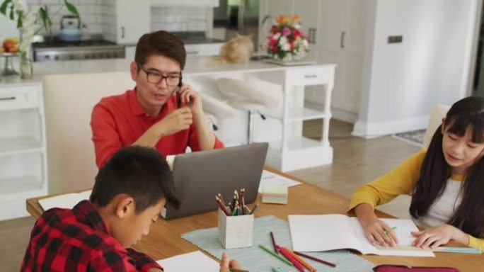 亚洲父亲使用智能手机和笔记本电脑在厨房工作，儿子和女儿在做功课