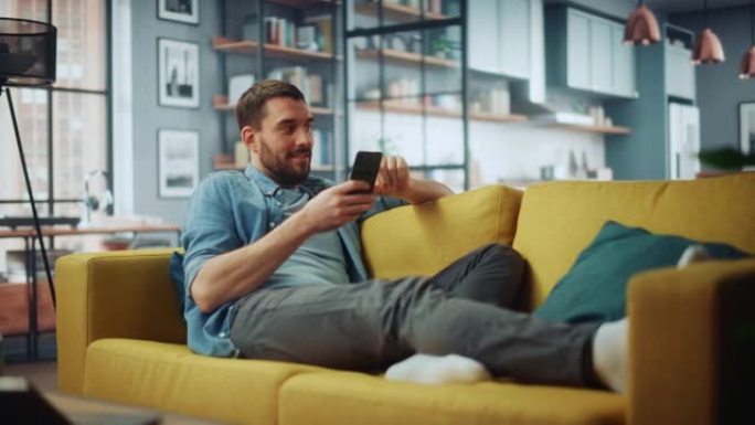 快乐英俊的白人男人在家里舒适的客厅使用智能手机。男人躺在舒适的沙发上。他正在浏览互联网，并在社交网络