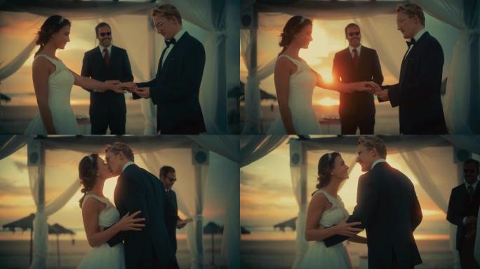 日落时分，在海洋海滩举行的户外婚礼上，新娘和新郎的特写镜头。浪漫情侣结婚、交换戒指、亲吻和与朋友分享