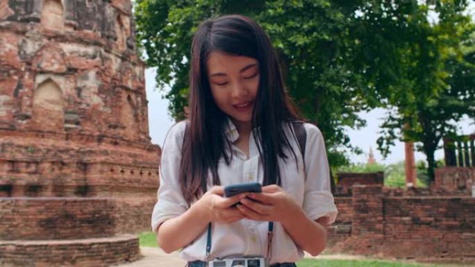 开朗美丽的年轻亚洲背包客博主女人休闲与相机看照片智能手机微笑与快乐的脸旅行宝塔在老城。