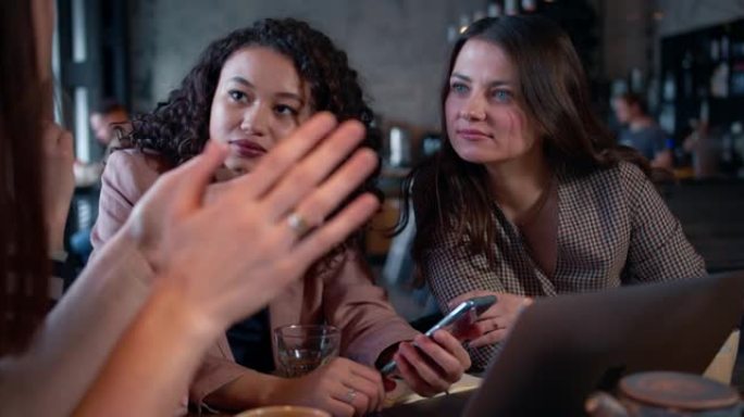 合作理念。快乐美丽的多元化商业女性在咖啡馆一起工作，聊天，使用智能手机和笔记本电脑。
