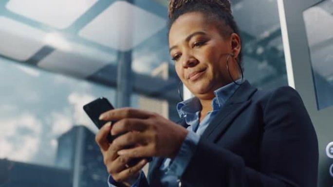 成功的黑人成年女性乘坐玻璃电梯到现代商务中心的办公室。美丽的非洲裔美国经理使用智能手机，写信息，查看