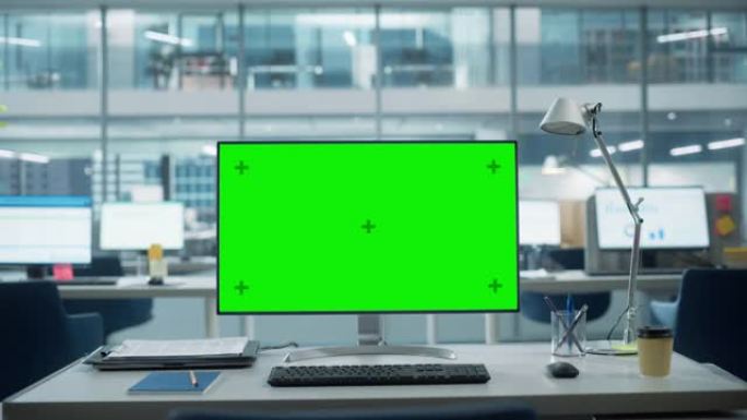 台式电脑显示器，带有模拟绿屏色度键显示屏，站在现代商务办公室的桌子上。在带有大城市办公室的背景玻璃墙