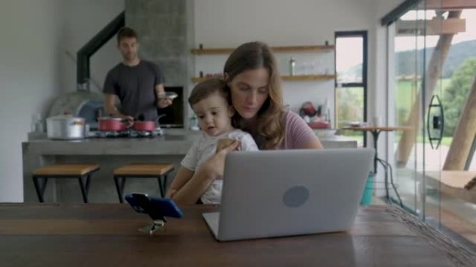 父亲为儿子和妻子准备午餐，而她在笔记本电脑上工作并抱着婴儿