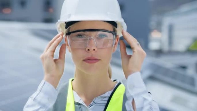 一名年轻的白人妇女在安装太阳能电池板时戴着护目镜和安全帽。一位自信的女员工站在建筑工地的建筑物上的肖