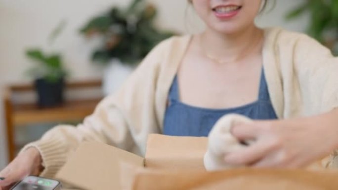 年轻的亚洲女性从她在家网上购物零售送货的纸板箱中拆箱新购买的衣服。