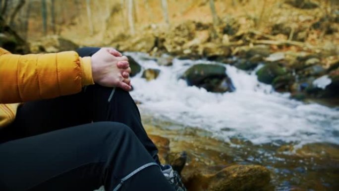 穿着暖和衣服的女人坐在森林流水旁边的岩石上