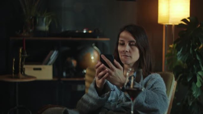 处理孤独。年轻迷人的悲伤女人使用智能手机社交媒体应用程序在黑暗的客厅喝酒。
