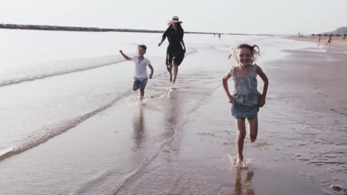 爱情和家庭度假，两个快乐的孩子在日出海滩水上与年轻的母亲慢动作一起玩耍。