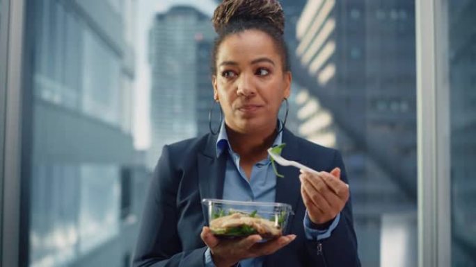 自信的非洲裔美国女性乘坐玻璃电梯到现代商务中心的办公室，吃健康的蔬菜沙拉。快乐的经理为工作中富有成效