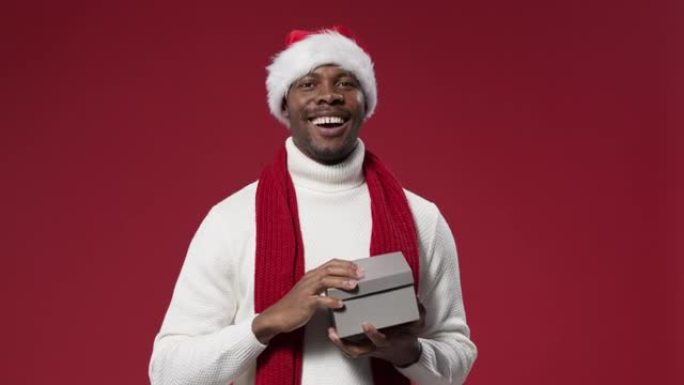 一个戴着圣诞帽和羊毛针织毛衣的黑皮肤男人拿了一个礼品盒，打开它，开心地笑着，眯着眼睛
