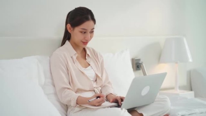 亚洲女商人在床上写记事本和使用技术笔记本电脑进行在线学习