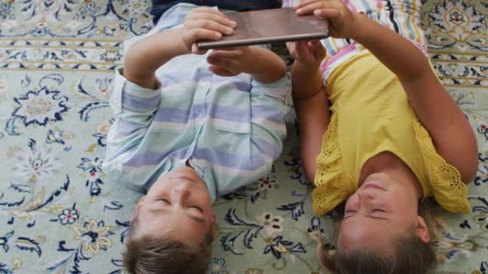 快乐的高加索兄弟姐妹在家，躺在地板上一起使用平板电脑，微笑着