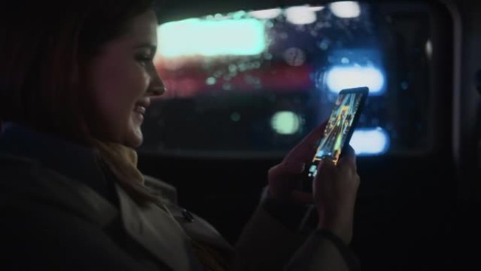 快乐的女性晚上坐出租车后座回家。美丽的女人乘客在智能手机上玩街机视频游戏，而在城市街道上的一辆有工作