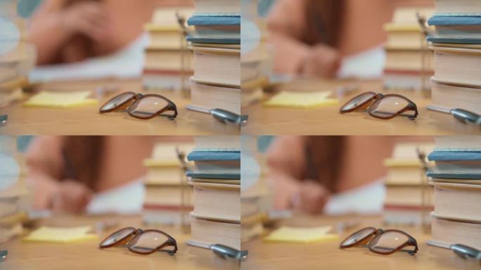 在图书馆或后台在家学习学术期末考试时，一副眼镜和一名学生写笔记的特写镜头。敬业的女人为她的教育做准备