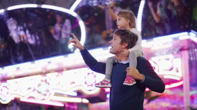 真实的照片，一个快乐的微笑的父亲在晚上带着露娜公园的灯光在游乐园里一起玩乐，肩上扛着他的小女儿。