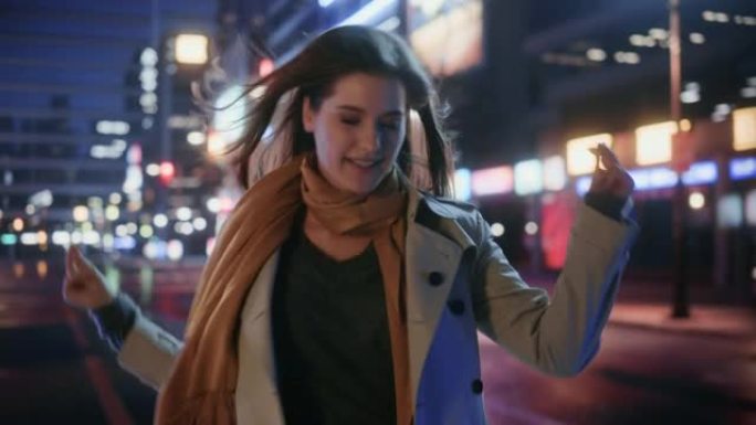 一个穿着风衣的幸福美女的肖像在夜晚霓虹灯的现代城市街道上行走。迷人的女性微笑着跳来跳去，兴奋地来到城