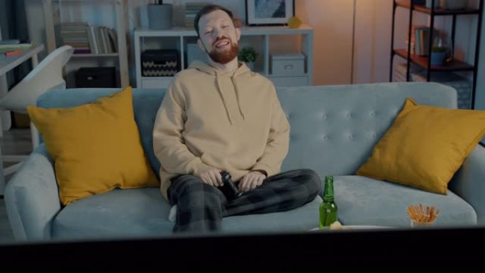 无忧无虑的家伙晚上在公寓里玩电子游戏和喝啤酒看电视屏幕的肖像