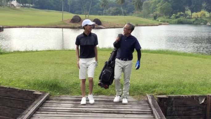 一名亚洲华裔男子带着他的十几岁的儿子走向高尔夫球场，带着高尔夫球袋看着风景