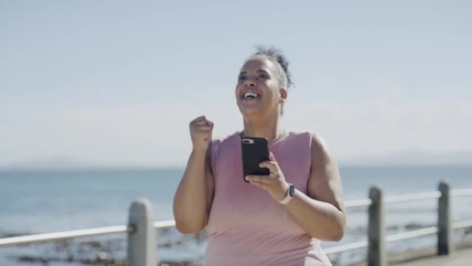 一位年轻的大码女士庆祝减肥之旅，并专注于健康，有氧运动，健身和精力。幸福的女人在海滩上的手机上安装了