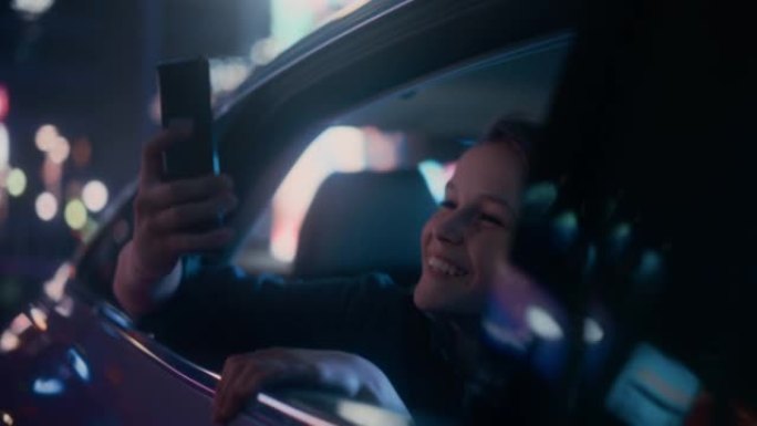 晚上，兴奋的小男孩从汽车后座望向窗外。孩子在智能手机上拍摄照片和视频，惊讶地发现带有工作霓虹灯的城市