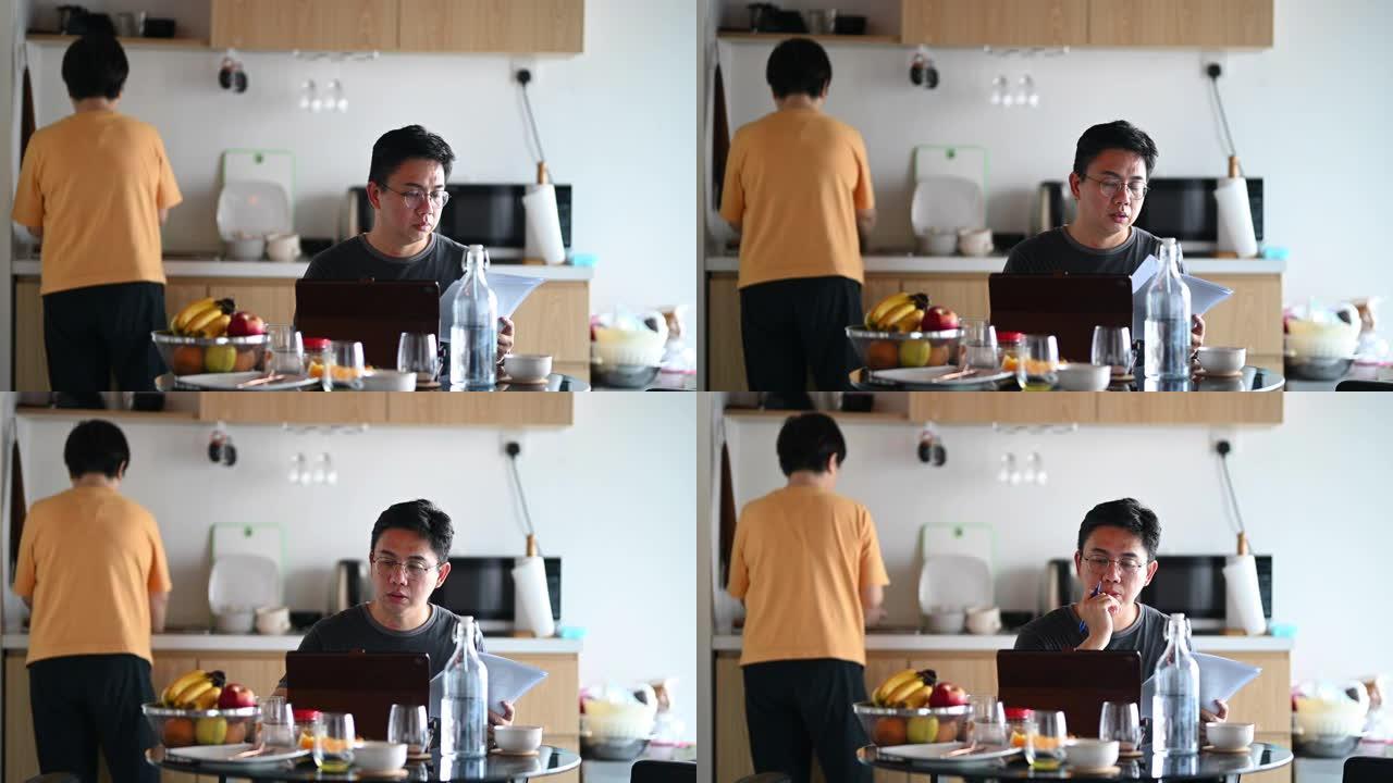 一名亚洲中国中年男子在早餐时间在家餐桌上工作，使用数字平板电脑和文件，妻子洗碗