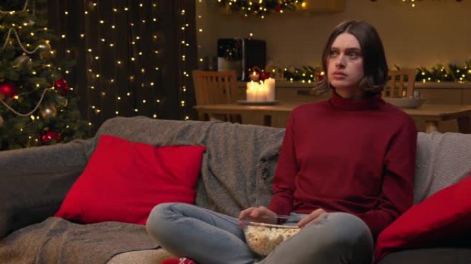 一个穿着红色毛衣的年轻美女坐在舒适的圣诞节装饰房间的沙发上，拿着一碗爆米花在电视上看恐怖电影