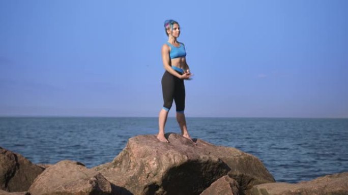 一位年轻漂亮的运动女子，有着绿松石般的头发，正站在鹅卵石上靠着大海和天空做运动，