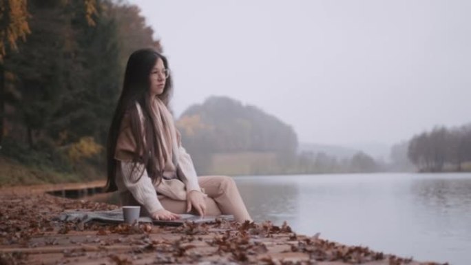 年轻女子坐在湖边的木板路上，关上笔记本电脑的盖子，拿起纸杯