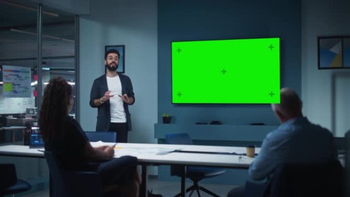 公司运营经理为一组经济学家举行会议演示。成年男性使用带有水平绿屏模拟显示的宽电视机。人们在商务办公室