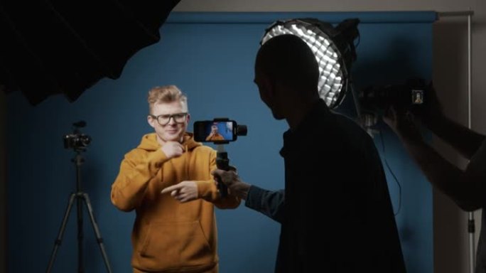 将手机放在稳定器中的操作员在带有蓝色背景的专业工作室中为一个受欢迎的视频博客拍摄视频