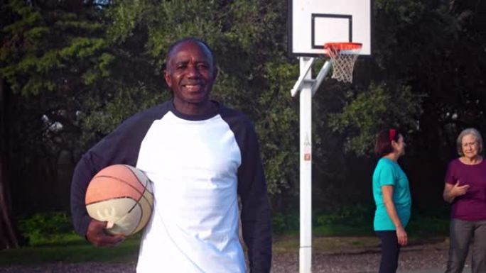 非裔美国高级男子站在户外球场上微笑着拿着篮球