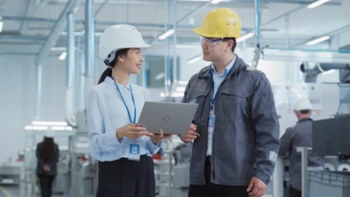 两名年轻的亚洲重工业工程师戴着安全帽的肖像，笔记本电脑在工厂里聊天。两名在生产工厂工作的制造员工的慢