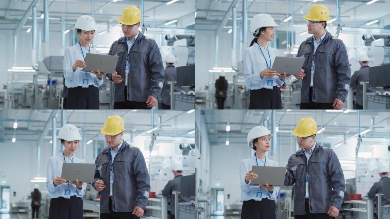 两名年轻的亚洲重工业工程师戴着安全帽的肖像，笔记本电脑在工厂里聊天。两名在生产工厂工作的制造员工的慢