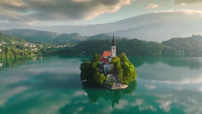 布莱德湖中心的圣母升天教堂的鸟瞰图。在斯洛文尼亚的布莱德湖上的小岛上飞行，温暖的早晨阳光和轻雾
